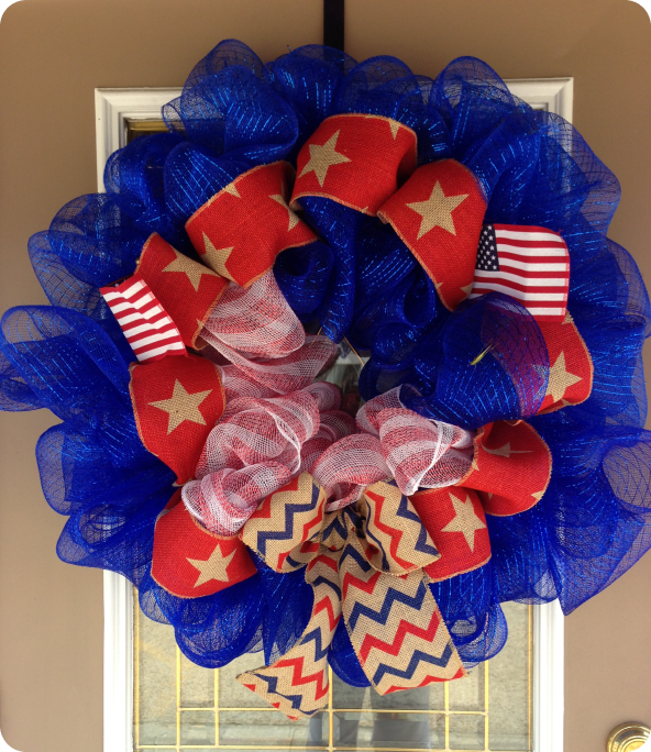 Patriotic wreath with blue mesh, red ribbon featuring gold stars, small American flags, white mesh accents, and a bow with red, blue, and tan chevron pattern, hanging on a door.