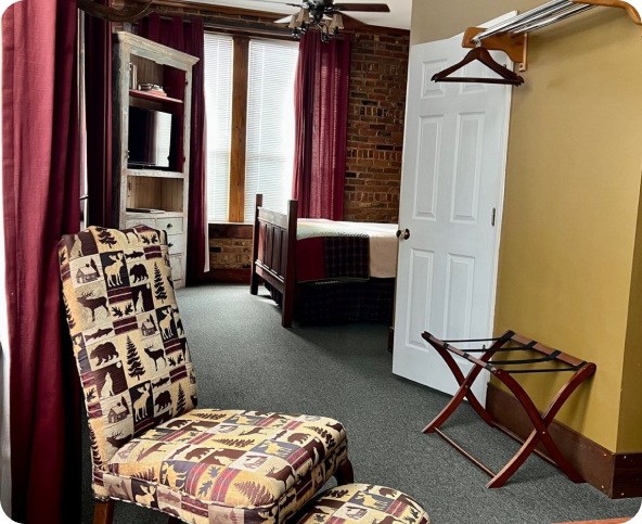 A cozy hotel room with rustic decor, featuring a patterned chair, a bed, brick accent wall, large window with maroon curtains, open closet with hangers, luggage rack, and a TV on a shelf.