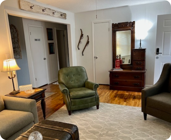 A cozy living room with green and gray armchairs, a beige sofa, a wooden chest, and a rug. An antique dresser with a mirror and vintage skis hung on the wall are visible. Warm lighting creates an inviting atmosphere.