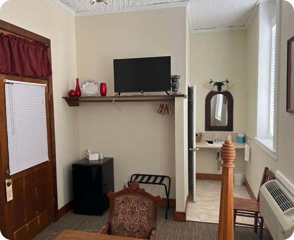 A small hotel room with a wooden door, a shelf holding a TV, decor items, a mini fridge, and a luggage rack. A chair and part of a bed are visible, with an open door revealing a bathroom sink and mirror.