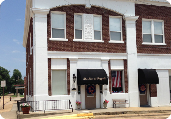 A red-brick building with white trim features two black awnings over doors, one reading “The Inn at Popeye.” An American flag hangs in a window, and potted flowers decorate the entrance steps.