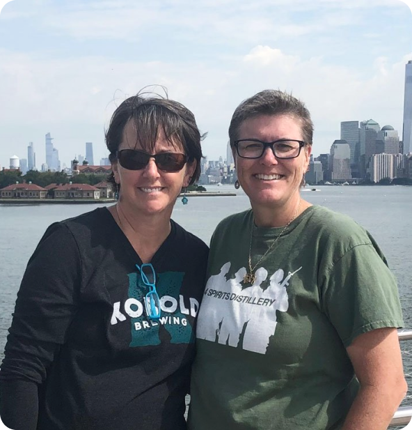 Two women smiling and standing close together outdoors with water and a city skyline, including tall buildings, in the background. Both are wearing sunglasses and casual t-shirts.