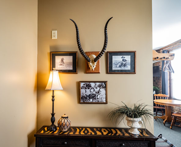 A room corner with a wall-mounted antelope skull, three framed black-and-white photos, a lit table lamp, pottery, a plant in a white pot, and a wooden table with a zebra-patterned runner.
