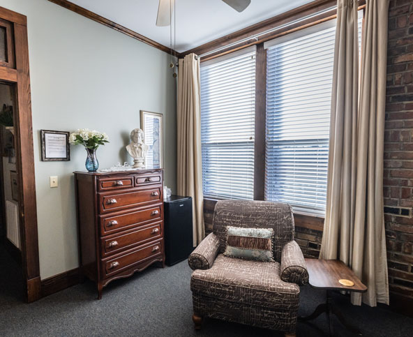 A cozy room with a patterned armchair, wooden side table, tall dresser with flowers and a bust, large curtained windows, and brick wall accents. Natural light fills the space.