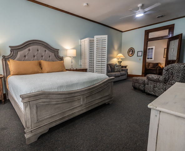 Spacious bedroom with a large wooden bed, two gold pillows, a white quilt, a patterned armchair, a sofa, lamps, a divider, and light blue walls with wood trim and carpeted floor.