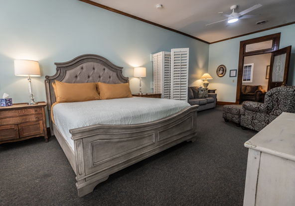A spacious bedroom with a large gray upholstered bed, two wooden nightstands with lamps, a sofa, an armchair, a dresser, and a folding room divider against pale blue walls and carpeted floor.