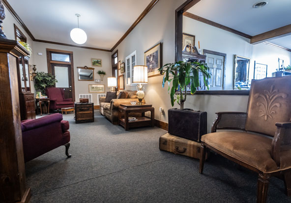 A cozy living room with vintage furniture, including armchairs, a sofa, wooden tables, potted plants, and framed art on beige walls, featuring warm lighting and a carpeted floor.