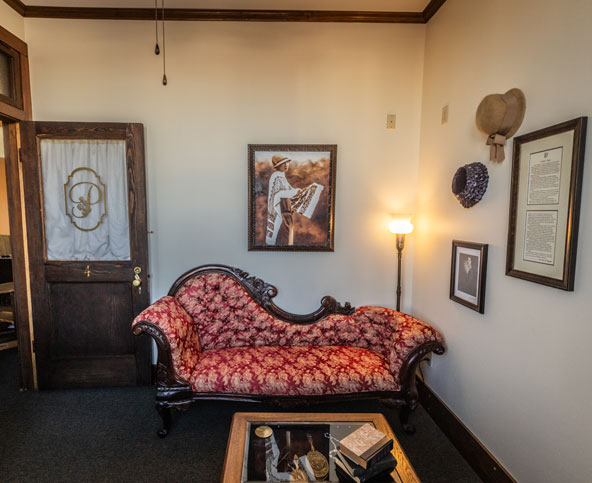 A vintage-style room with a red patterned chaise lounge, a framed portrait on the wall, hats hanging, a glass coffee table with books, and a wooden door with frosted glass and a monogram.