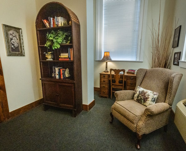 A cozy room with an upholstered armchair, a floral pillow, a wooden bookshelf with books and a plant, a small desk with a chair, a lamp, framed art, and a window with blinds.