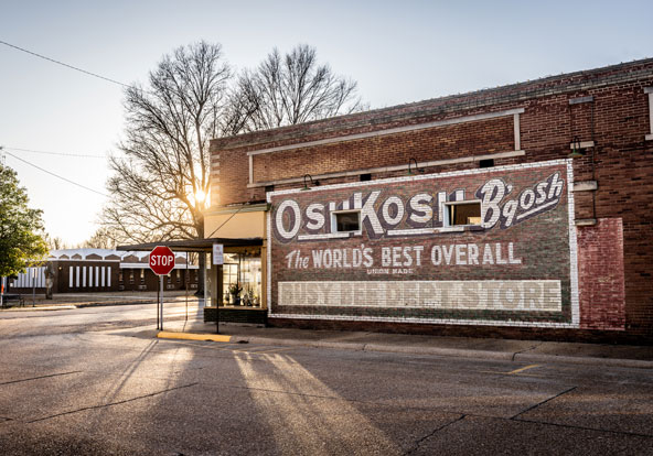 A brick building with a large vintage OshKosh B'gosh advertisement painted on the side, a stop sign at the corner, and sunlight shining through trees in the background.