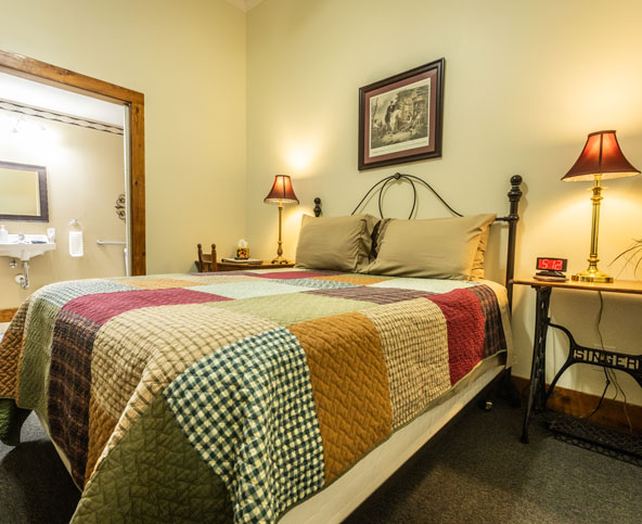 A cozy bedroom with a patchwork quilt on a double bed, two pillows, bedside tables with lamps, a framed picture above the bed, a clock reading 5:12, and an open door leading to a bathroom.