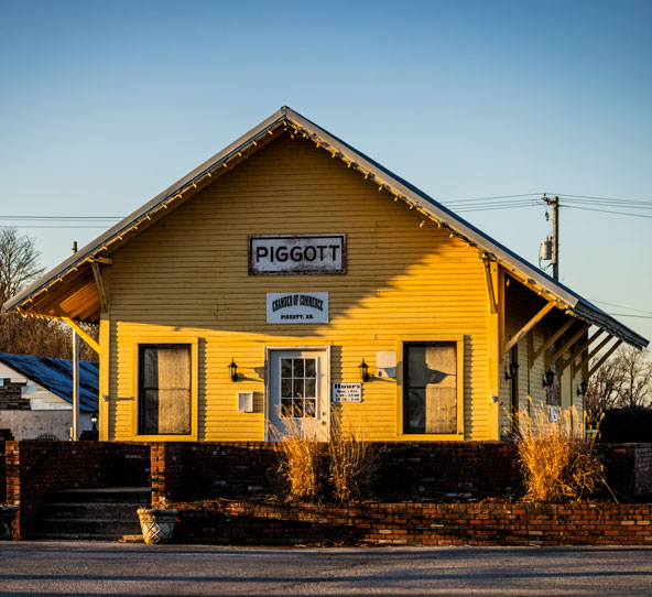A yellow wooden building with a sign reading "Piggott" and "Chamber of Commerce" above the entrance, surrounded by dry grass and bathed in golden sunlight.