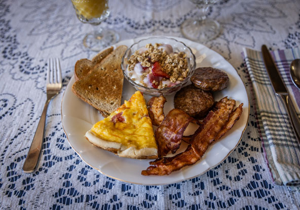 A breakfast plate with toast, a slice of egg casserole, crispy bacon, sausage patties, and a bowl of yogurt with granola and strawberries, set on a lace tablecloth with utensils and a glass of juice in the background.