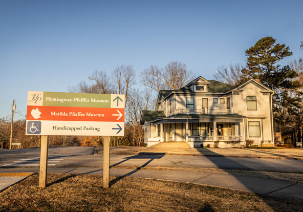 A large, historic white house with green roof sits behind a sign directing visitors to the Hemingway-Pfeiffer Museum, Matilda Pfeiffer Museum, and handicapped parking under a clear sky.