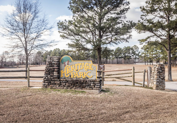 A large stone sign reading “Heritage Park” stands at the entrance to a grassy, open park area, surrounded by a split-rail wooden fence and tall pine trees under a partly cloudy sky.
