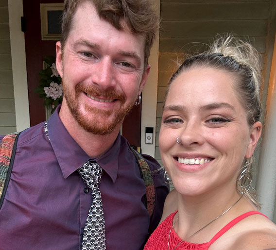 A smiling man and woman pose for a selfie outside a house. The man has short brown hair, a beard, and wears a purple shirt with a patterned tie. The woman has blonde hair in a bun and wears a red top with hoop earrings and a nose ring.